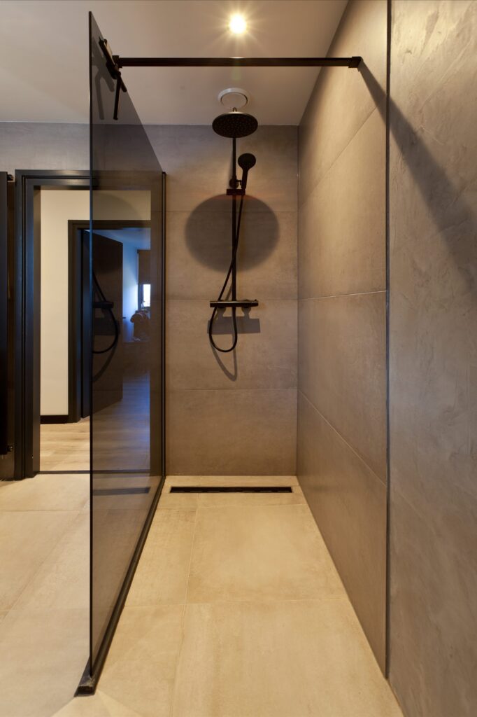 Inloopdouche met glazen wand met zwart profiel en grote metallook wandtegels in een badkamer in Rijnsburg