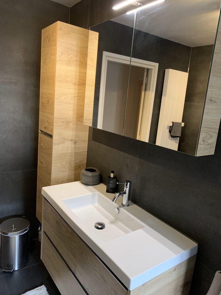 Badkamer in Katwijk met donkere matte wandtegels, wastafelmeubel en hoge kast in houtlook