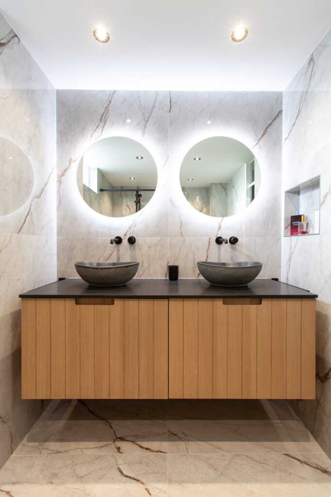 Badkamer in Leiderdorp met lichte marmerlook tegels, een houten badmeubel en twee ronde spiegels