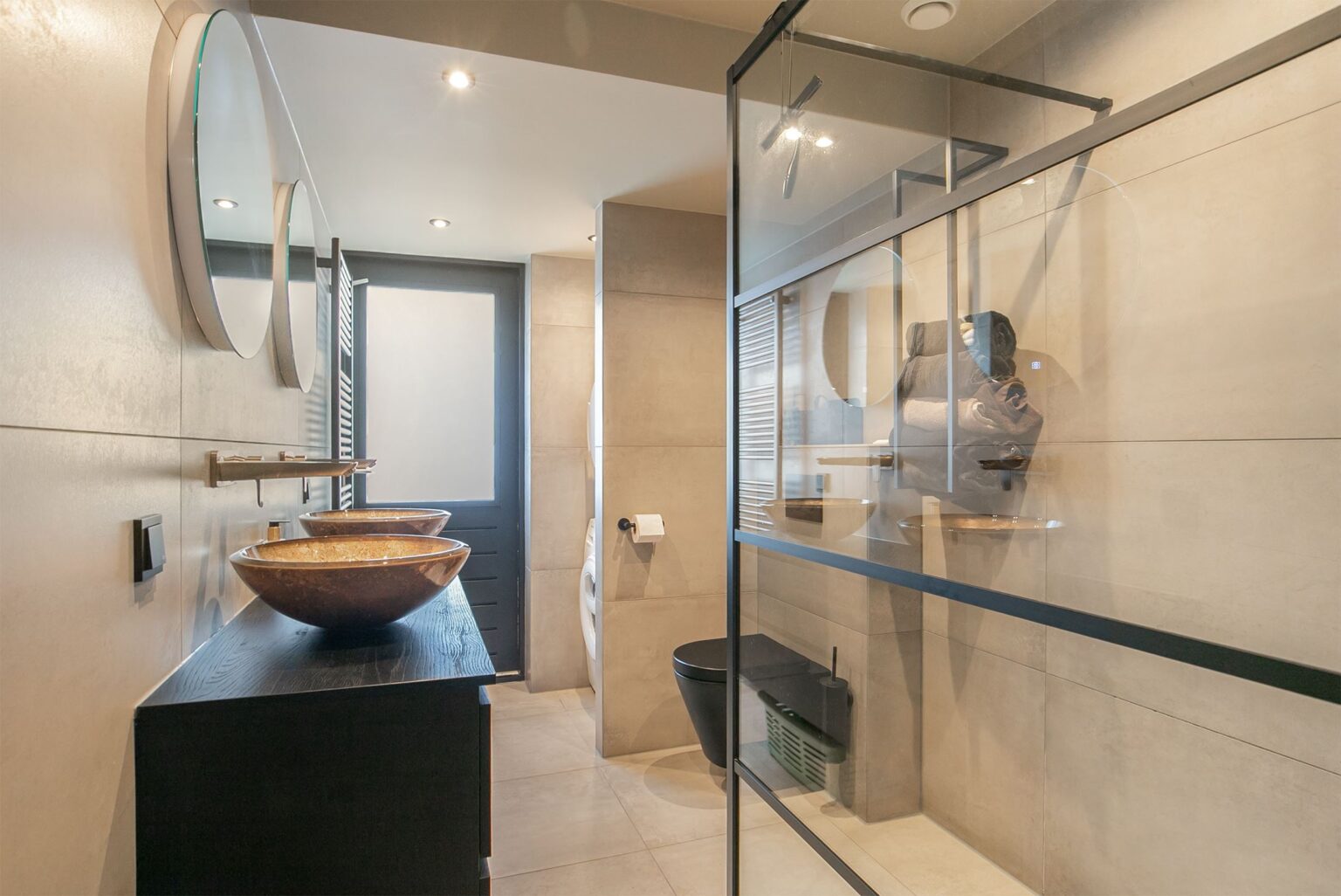Badkamer in Katwijk met lichte tegels, een donker badmeubel met koperen kommen en een glazen douchewand