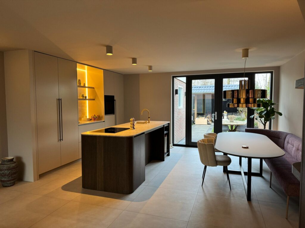 Woonkeuken in Sassenheim met zandgrijze betonlook vloertegels van 90×90, een kookeiland en een eethoek bij de tuindeuren