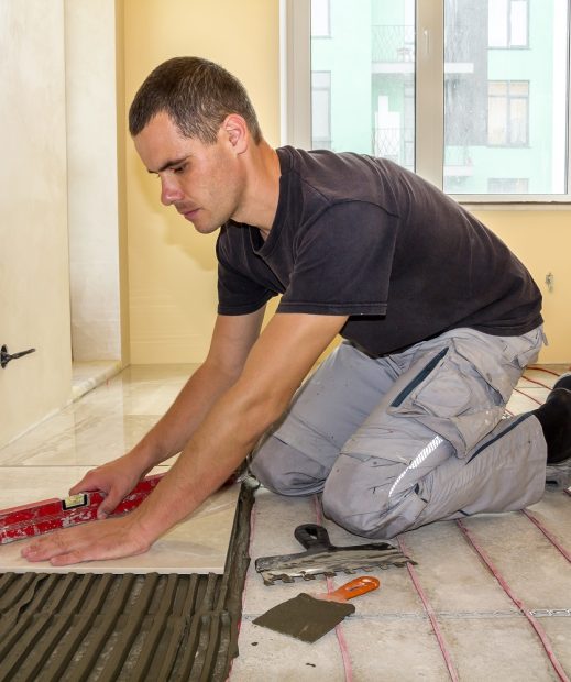 Young worker tiler installing ceramic tiles using lever on cement floor with heating red electrical cable wire system. Home improvement, renovation and construction, comfortable warm home concept.